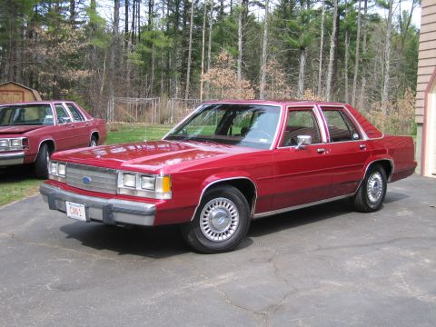 1991 Ford LTD Crown Victoria - Two red Ford Crown Victoria sedans are parked on a driveway with trees in the background.