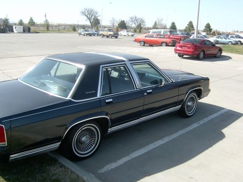 1990 Ford Crown Victoria - A black sedan with chrome accents and wire wheels is parked in a lot with other vehicles.