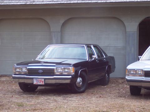 2001 Ford Crown Victoria - A dark sedan is parked in front of a garage, with another vehicle partially visible to the right.