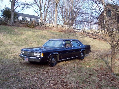 2001 Ford Crown Victoria - Dark sedan parked on a grassy slope near a stone wall and house.