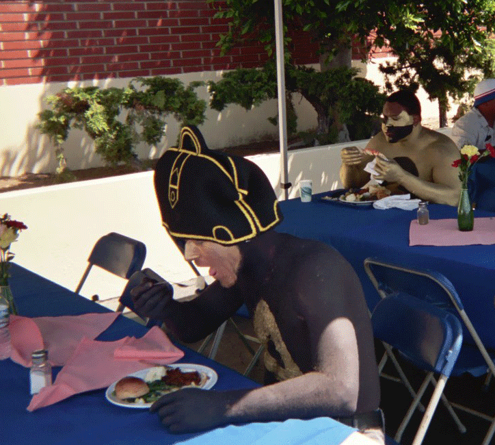 People in black bodysuits and dog masks eating at outdoor tables with blue cloths