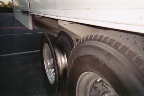 1990 Ford Bronco - shredded blown-out tire on the tandem dual axle of a white trailer or RV parked on asphalt at dusk.
