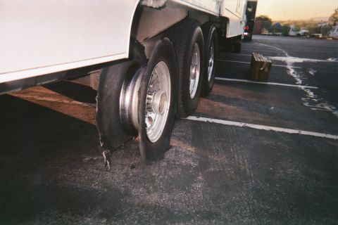 1990 Ford Bronco - completely destroyed tire with bare rim exposed on the tandem axle of a white trailer at dusk in a parking lot.