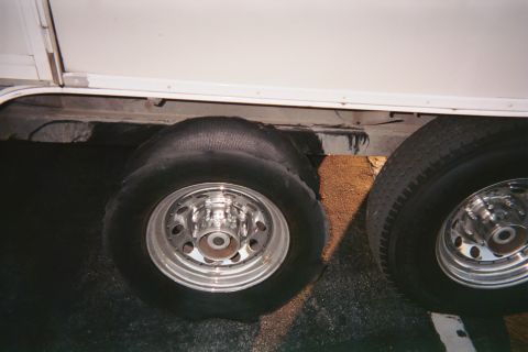 1990 Ford Bronco - flat, shredded tire on a chrome dually-style wheel under a white trailer, with a second wheel visible alongside.