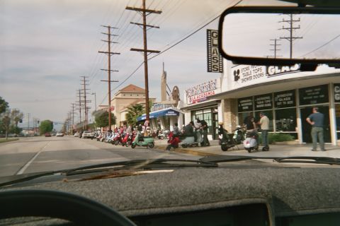 1968 Ford Mustang - A line of scooters is parked outside shops on a sunny day.