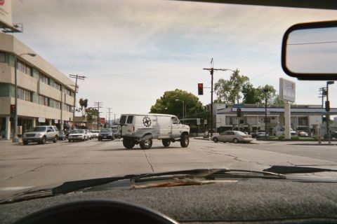 1990 Ford Bronco - Seen through a windshield, a white lifted van with a star door logo crosses a wide city intersection past low buildings and a gas station.
