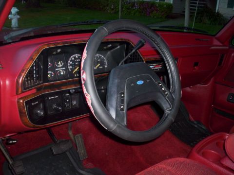 1989 Ford Bronco - The interior of a red Ford vehicle features a wood-grain dashboard and a black steering wheel with a prominent Ford emblem.
