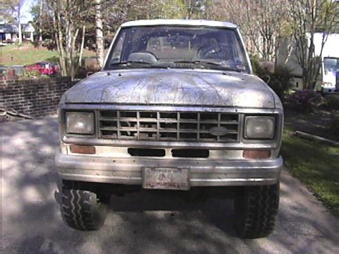 1984 Ford Bronco II - A dusty 1984 Ford Bronco II is shown from the front, parked outdoors with trees and houses in the background.
