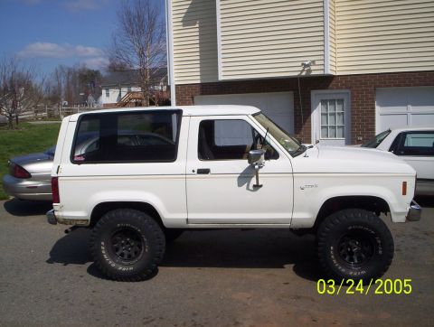 1984 Ford Bronco II - A white 1984 Ford Bronco II is parked on a street next to a house and another vehicle.