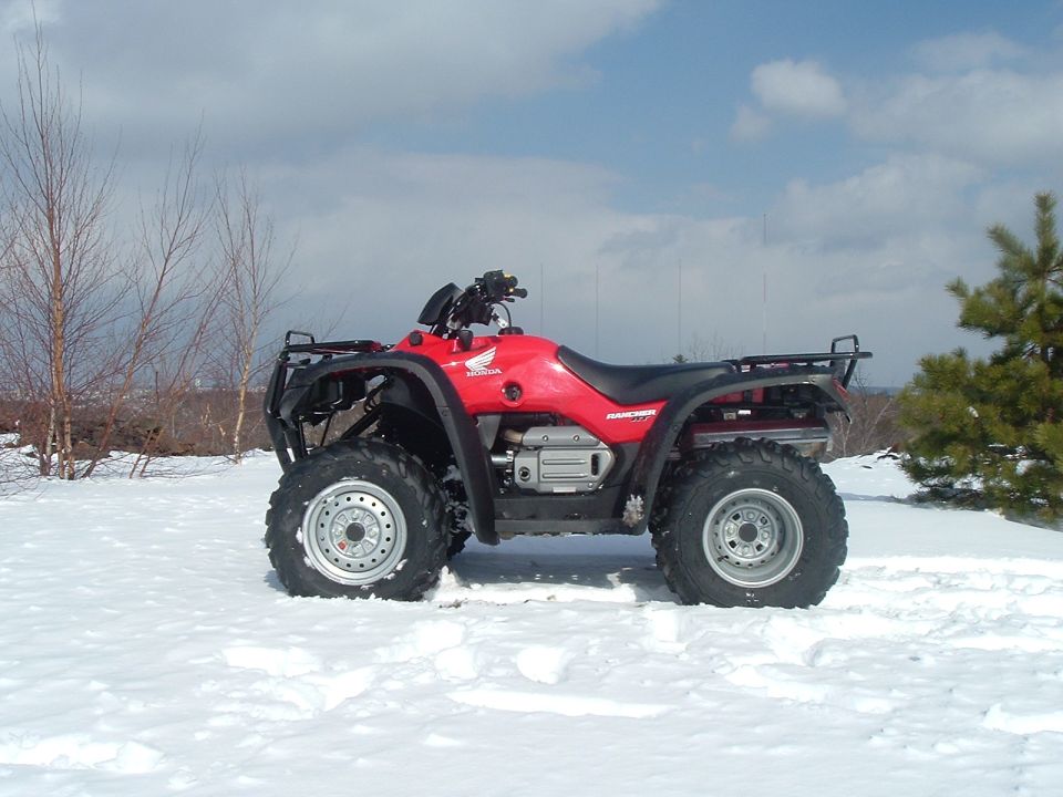 Red Honda Rancher ATV parked on snow with bare trees and cloudy sky