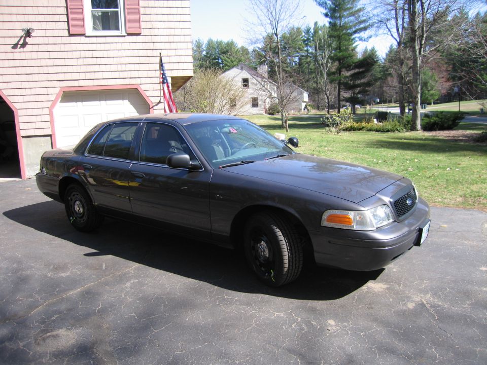 Dark gray Ford Crown Victoria parked in a driveway. It has black steel wheels and a police-style spotlight on the driver's side mirror