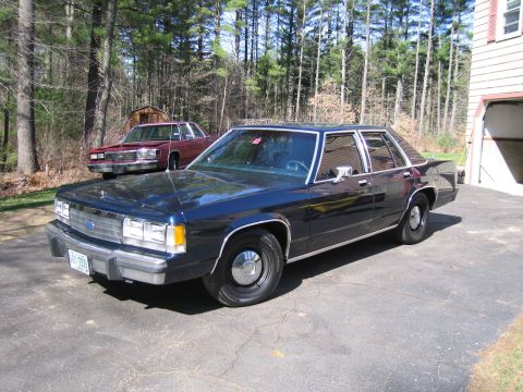 1991 Ford LTD Crown Victoria - A dark blue sedan is parked in a driveway with a maroon sedan behind it. The scene is set against a backdrop of trees and a house.