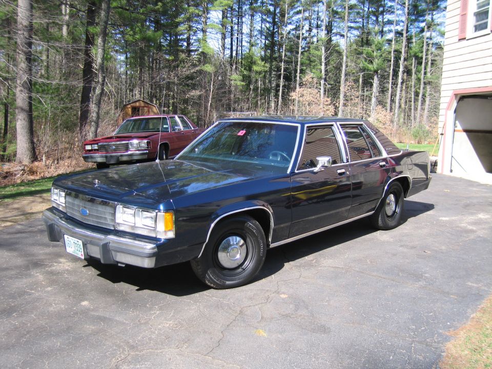 Dark blue sedan parked in driveway, maroon sedan behind it. Trees and house in background