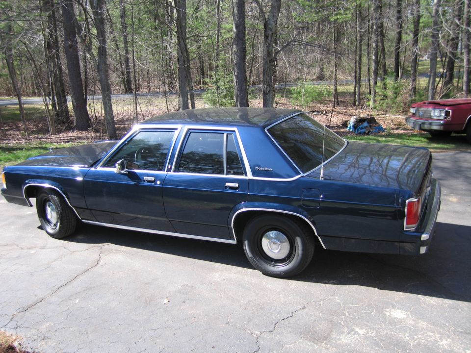 1991 Ford Crown Victoria LTD sedan, dark blue, parked on cracked asphalt driveway with trees in background. Red vehicle visible to the right
