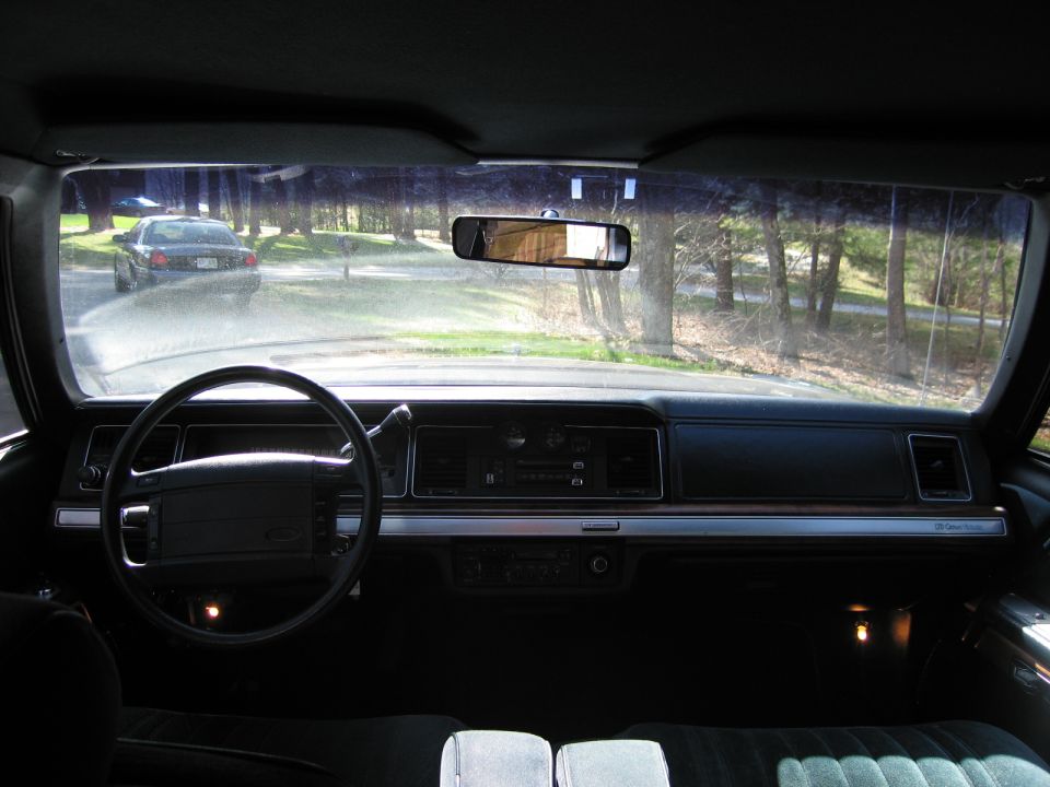 Interior view of a 1990s sedan showing dashboard, steering wheel, and a dark sedan outside through the windshield