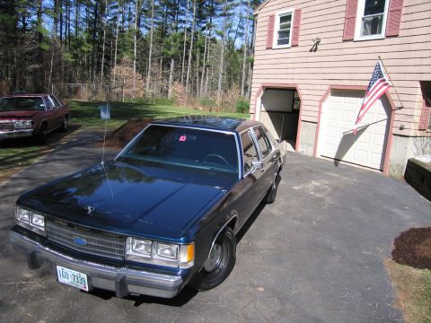 1991 Ford LTD Crown Victoria - A dark blue sedan is parked in a driveway next to a house with an American flag displayed.