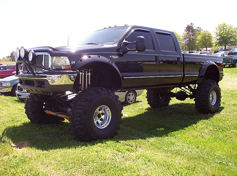 Lifted black pickup truck with big tires and extra lights on the front. Other vehicles are parked in the background