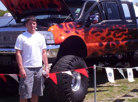1995 Ford Bronco - A man stands beside a heavily modified black truck with custom flame paint and oversized tires.