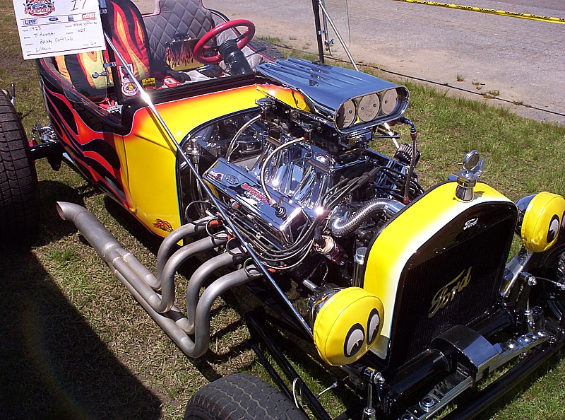 Custom hot rod with yellow and flame paint, exposed V8 engine, and large exhaust pipes. Ford logo on grille
