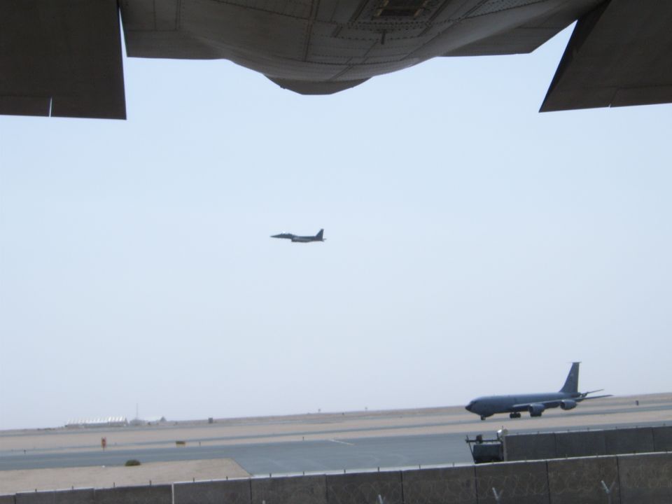 View from under an aircraft wing shows a tanker plane on the tarmac and a fighter jet in the sky