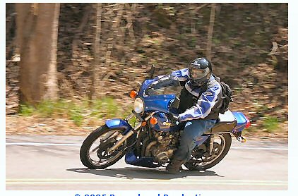 1985 Ford LTD Crown Victoria - A rider leans into a turn on a blue motorcycle on a tree-lined road.