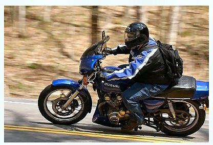 1985 Ford LTD Crown Victoria - A rider on a blue Suzuki motorcycle navigates a road, with trees blurred in the background.