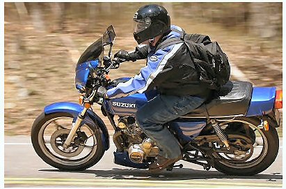 1985 Ford LTD Crown Victoria - Rider on a blue Suzuki motorcycle on a road, with trees in the background.