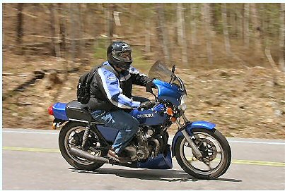 1985 Ford LTD Crown Victoria - A rider on a blue Suzuki motorcycle cruises down a tree-lined road.