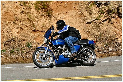 1985 Ford LTD Crown Victoria - Rider on a blue motorcycle navigates a road with a rocky embankment on the side.