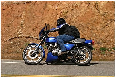 1985 Ford LTD Crown Victoria - A rider on a blue Suzuki motorcycle cruises down a paved road with a rocky embankment to their side.