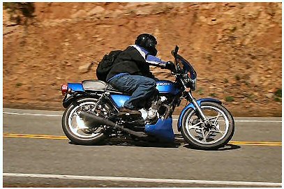 1985 Ford LTD Crown Victoria - A rider on a blue motorcycle navigates a road with a rocky hillside in the background.