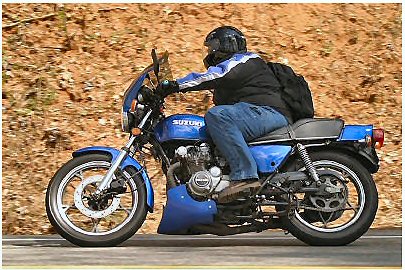 1985 Ford LTD Crown Victoria - A rider on a blue Suzuki motorcycle cruises down a road with a textured embankment in the background.