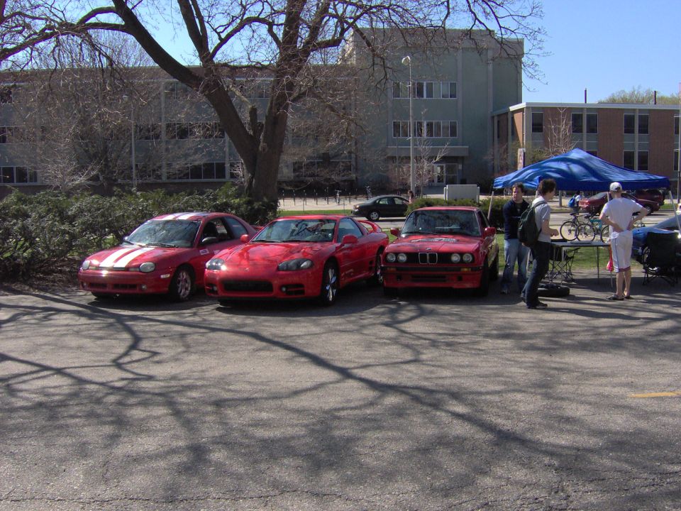Three red cars parked in a lot at a car meet with a blue canopy and people nearby
