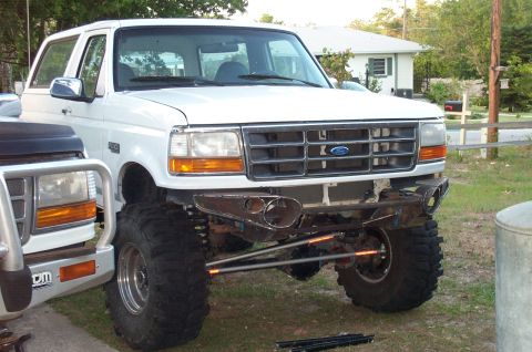 1995 Ford Bronco - A white lifted Ford Bronco sits in a yard mid-build with its front bumper removed, exposing the frame horns, solid front axle, crossover st…