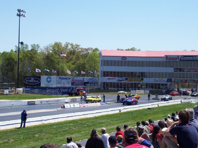 Drag strip with multiple cars, spectators, and a grandstand building