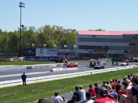 1978 Ford F-250 - Cars race down a drag strip with spectators watching from the stands and a large building behind the track.