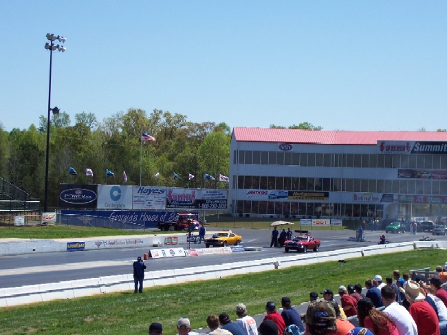 Drag strip with multiple cars, grandstands, and a multi-story building. Watermark: SUPER MOTORS www.supermotors.net