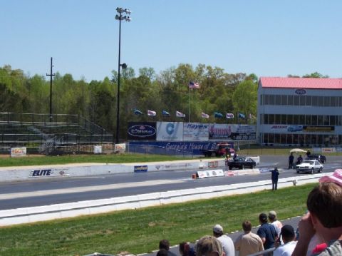 1978 Ford F-250 - Cars compete on a drag strip under a clear blue sky, with spectators in the foreground and a large building in the background.