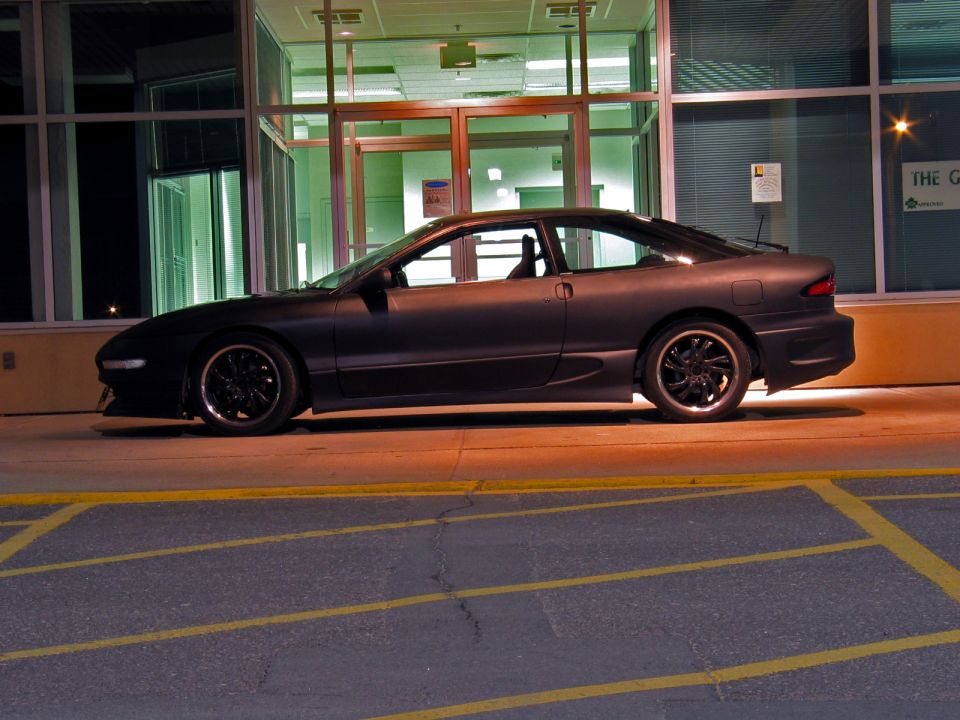 Matte black Ford Probe coupe in side view, parked at night outside a glass-front building