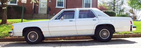 1985 Ford Crown Victoria LTD - A white sedan is parked on the side of the road in front of a brick house.