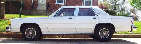 1985 Ford Crown Victoria LTD - This white sedan, likely from the 1980s, is parked curbside in front of a brick house.