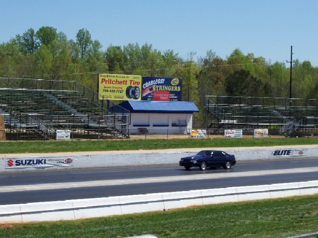 Dark blue sedan driving on a drag strip with bleachers and advertising signs in the background