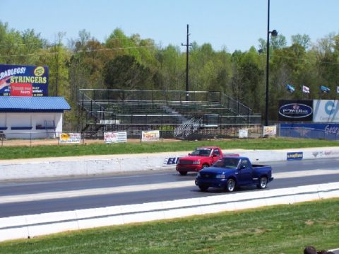 1978 Ford F-250 - Two pickup trucks compete on a drag strip, with spectators in the stands watching the race.