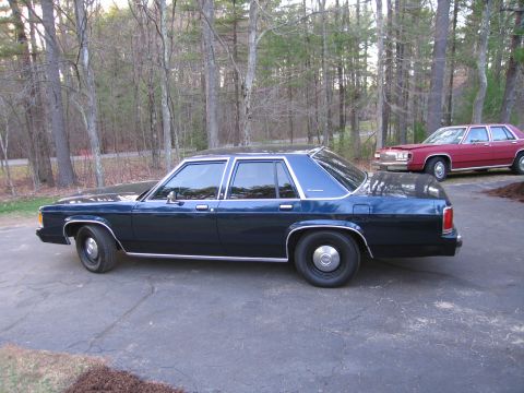 1991 Ford LTD Crown Victoria - A dark blue sedan is parked on an asphalt driveway with trees in the background.