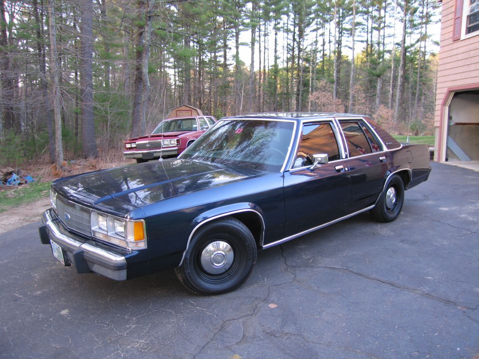 Dark blue sedan parked on asphalt driveway, another red sedan in background. Trees and house visible