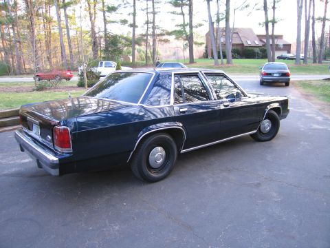1991 Ford LTD Crown Victoria - A dark blue sedan is parked on a paved driveway with trees and houses in the background.