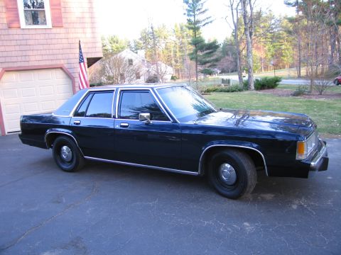 1991 Ford LTD Crown Victoria - A dark blue sedan, likely from the 1980s, is parked in a driveway next to a garage.