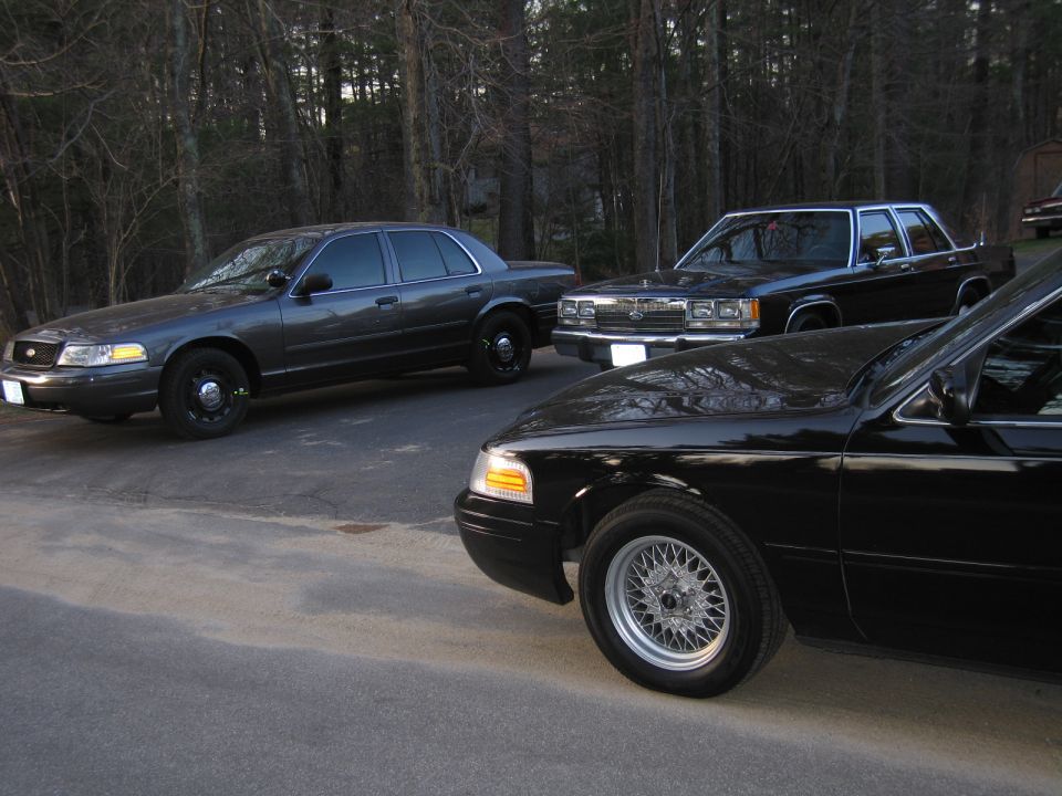 Three Ford Crown Victorias parked on a paved surface with trees in the background. One is dark gray, one is black, and one is partially visible in the foreground, also black