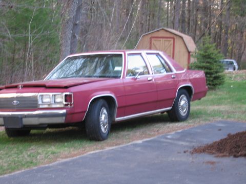 1991 Ford LTD Crown Victoria - red 1980s Ford LTD Crown Victoria sedan with a hood bra parked on a driveway near a wooden shed and bare trees.