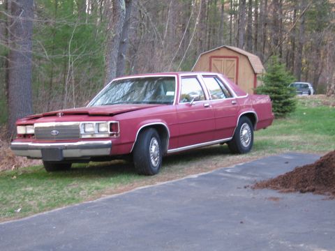 1991 Ford LTD Crown Victoria - Maroon sedan parked on grass with a shed and trees in the background.